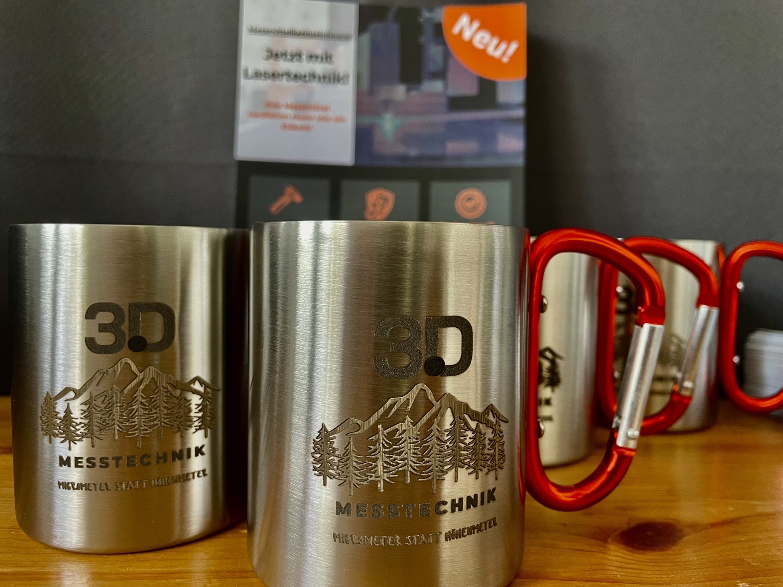 Fertige Edelstahlbecher mit Faserlaser-Gravur: 3D-Messtechnik-Firmenlogo mit Berg- und Tannenmotiv, Slogan 'MIKROMETER STATT H&Ouml;HENMETER' &ndash; Beispiel f&uuml;r individualisierte Werbeartikel und Geschenke.