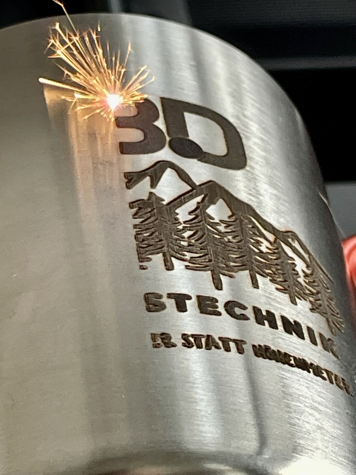 Live-Aufnahme einer Faserlaser-Gravur: Funkenflug w&auml;hrend der Lasermarkierung des 3D-Messtechnik-Logos auf einem Edelstahlbecher. Pr&auml;zise Tiefengravur in Aktion.